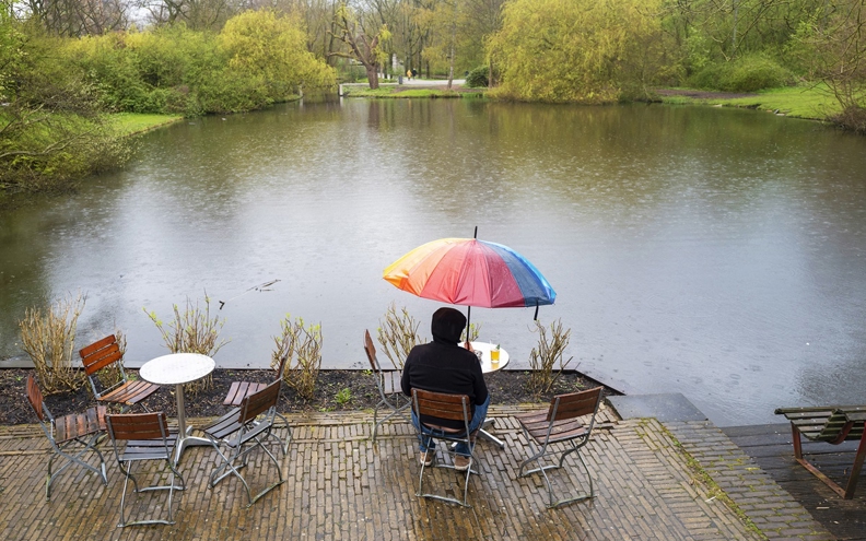 Man alleen in de regen onder een regenboog-paraplu op het terras aan de rand van het water