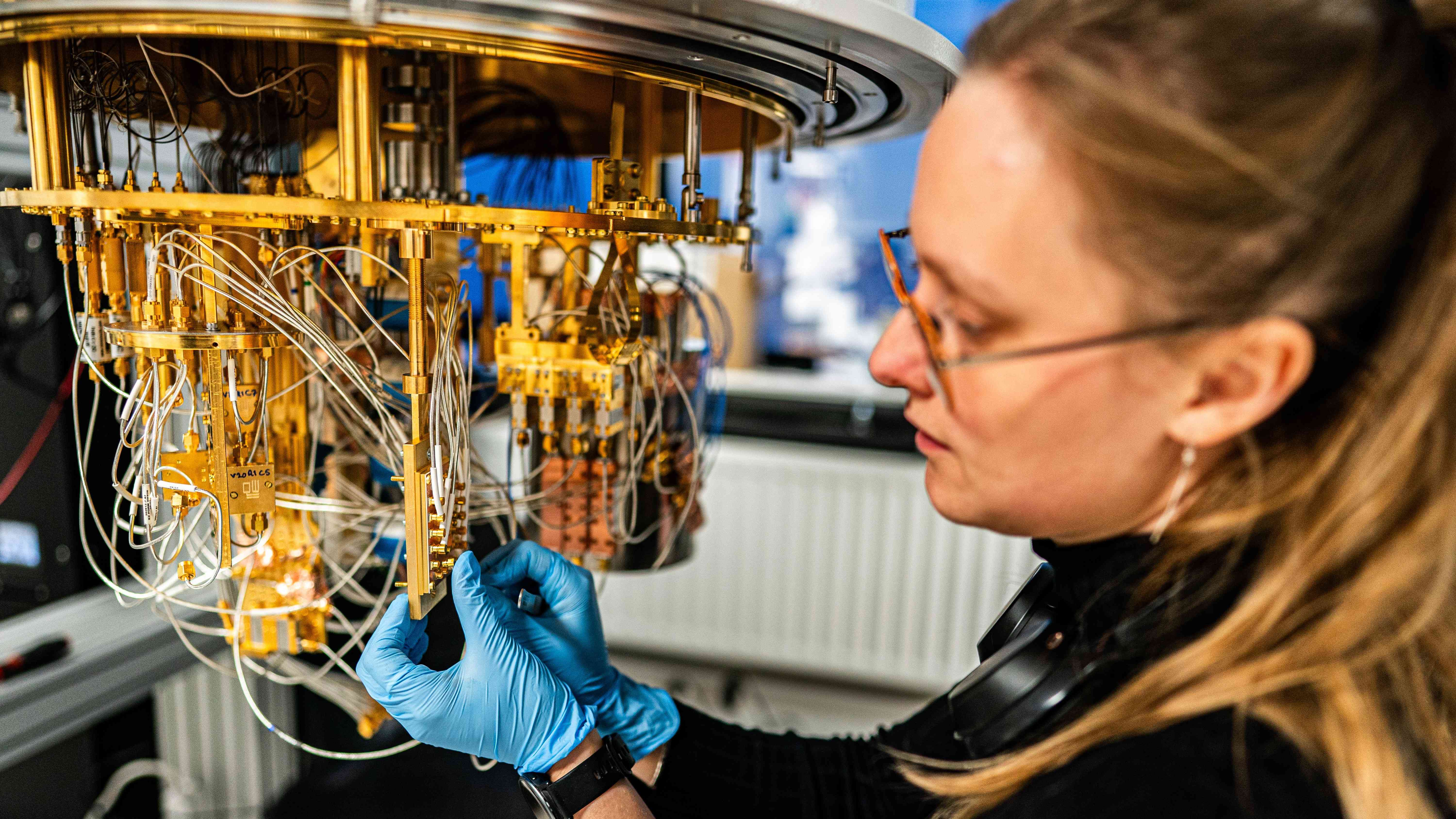 Een vrouw van ongeveer 35 jaar oud met blauwe wegwerphandschoenen werkt in een laboratorium aan een grote, hangende goudkleurige quantumprocessor met veel dunne kabels en kleine onderdelen. De persoon houdt een onderdeel van de installatie vast terwijl de draden naar beneden hangen. Op de achtergrond zijn laboratoriumapparatuur en een radiator zichtbaar.