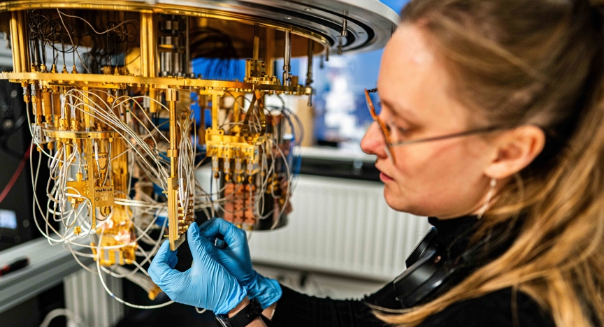 Een vrouw van ongeveer 35 jaar oud met blauwe wegwerphandschoenen werkt in een laboratorium aan een grote, hangende goudkleurige quantumprocessor met veel dunne kabels en kleine onderdelen. De persoon houdt een onderdeel van de installatie vast terwijl de draden naar beneden hangen. Op de achtergrond zijn laboratoriumapparatuur en een radiator zichtbaar.