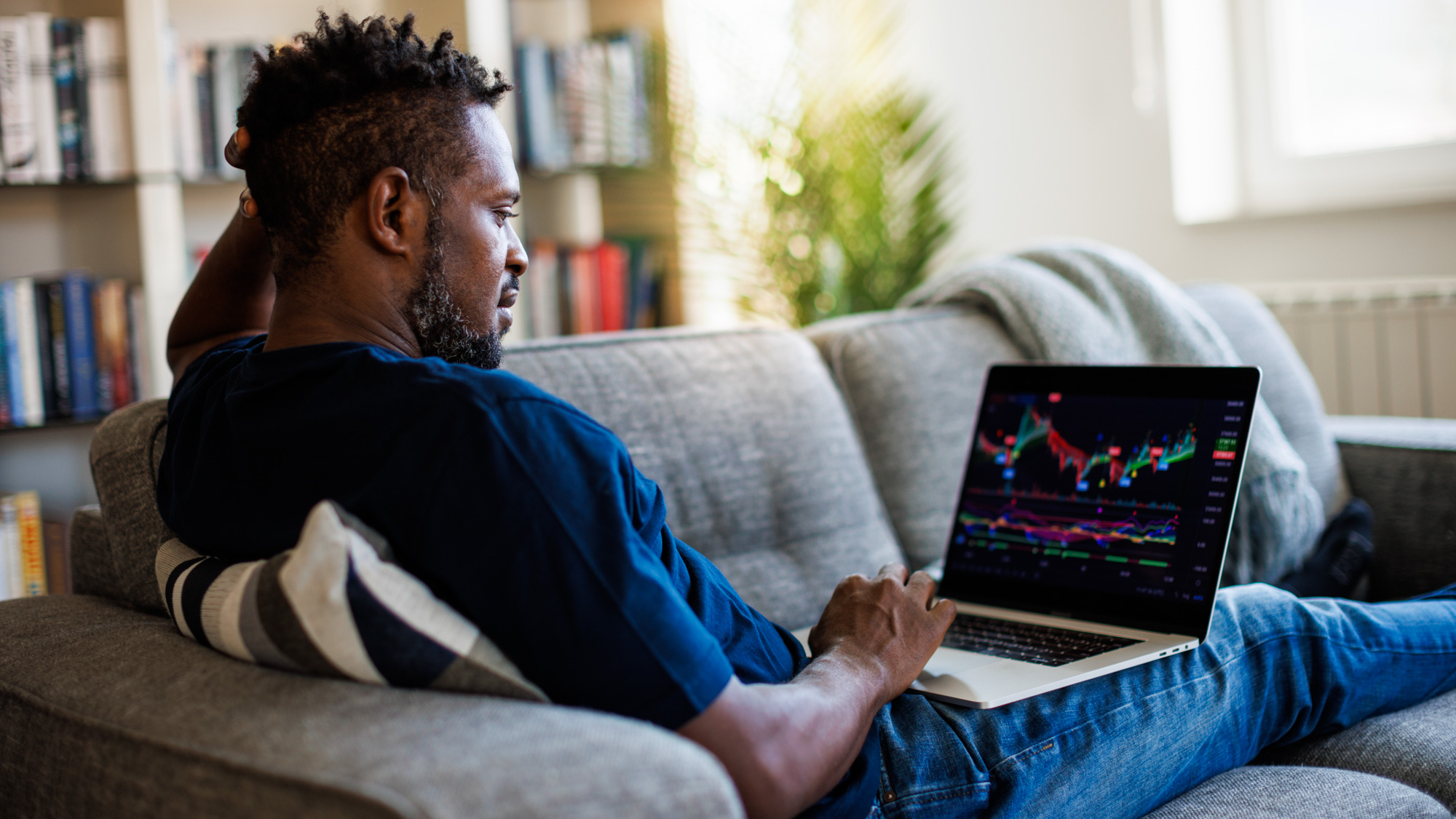 Man ligt op de bank met zijn laptop terwijl hij naar de koers van crypto valuta kijkt.
