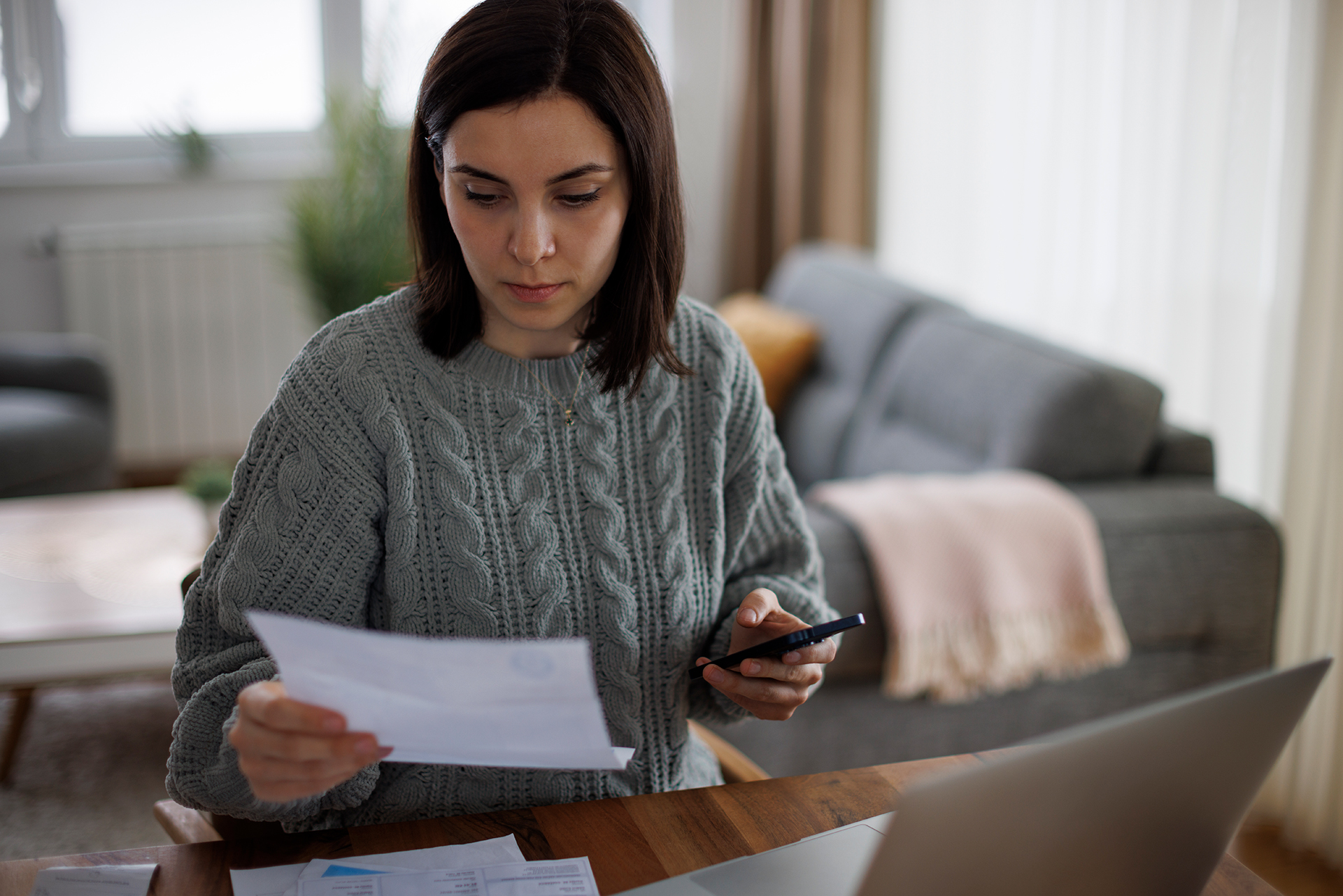 Persoon bekijkt een document en houdt een smartphone vast aan een tafel met laptop en papieren.