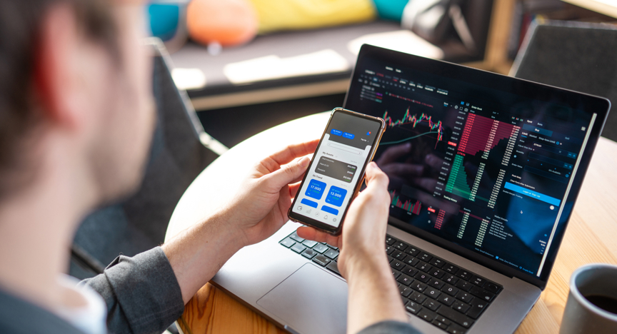 Een man kijkt naar zijn crypto investeringen op zijn smartphone en laptop.