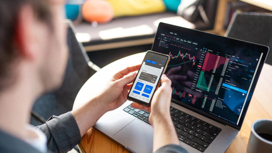 Een man kijkt naar zijn crypto investeringen op zijn smartphone en laptop.