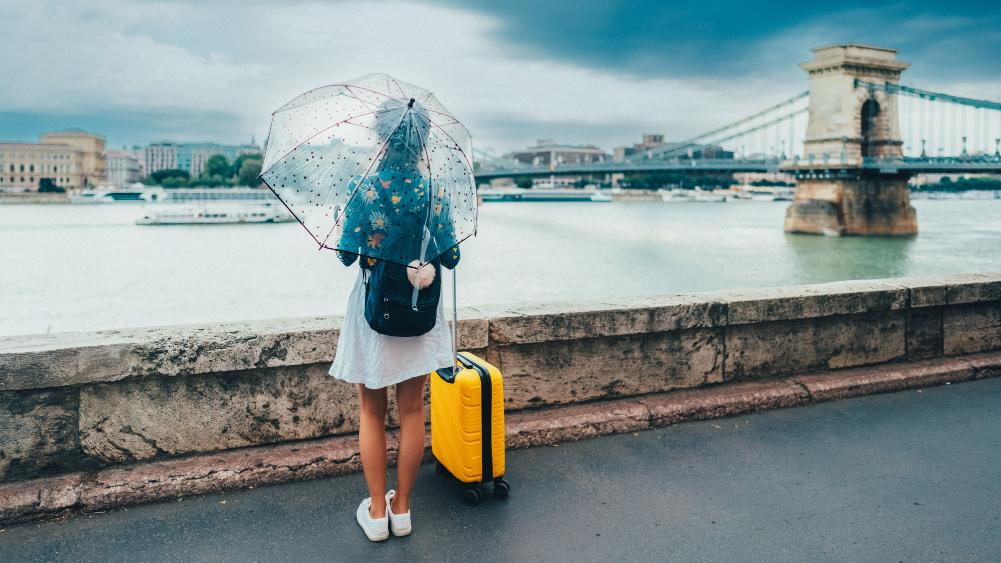 Een vrouw staat in zomerkleding, maar met paraplu, aan de rand van een rivier in een Europese stad en kijkt naar het uitzicht.