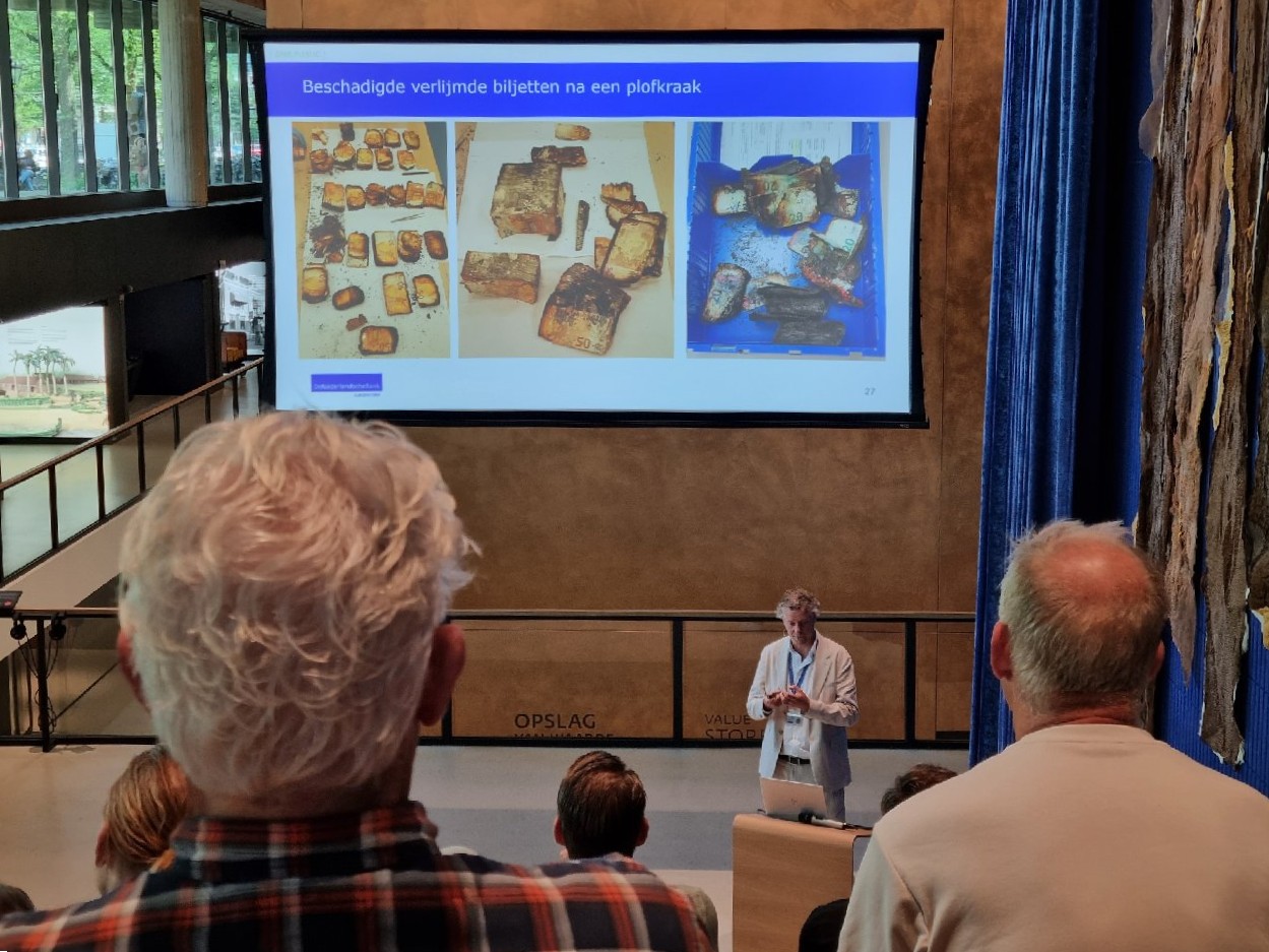 Lunchsessie bij DNB over plofkraken, mensen in de zaal en een slide met daarop een afbeelding van beschadigde biljetten.