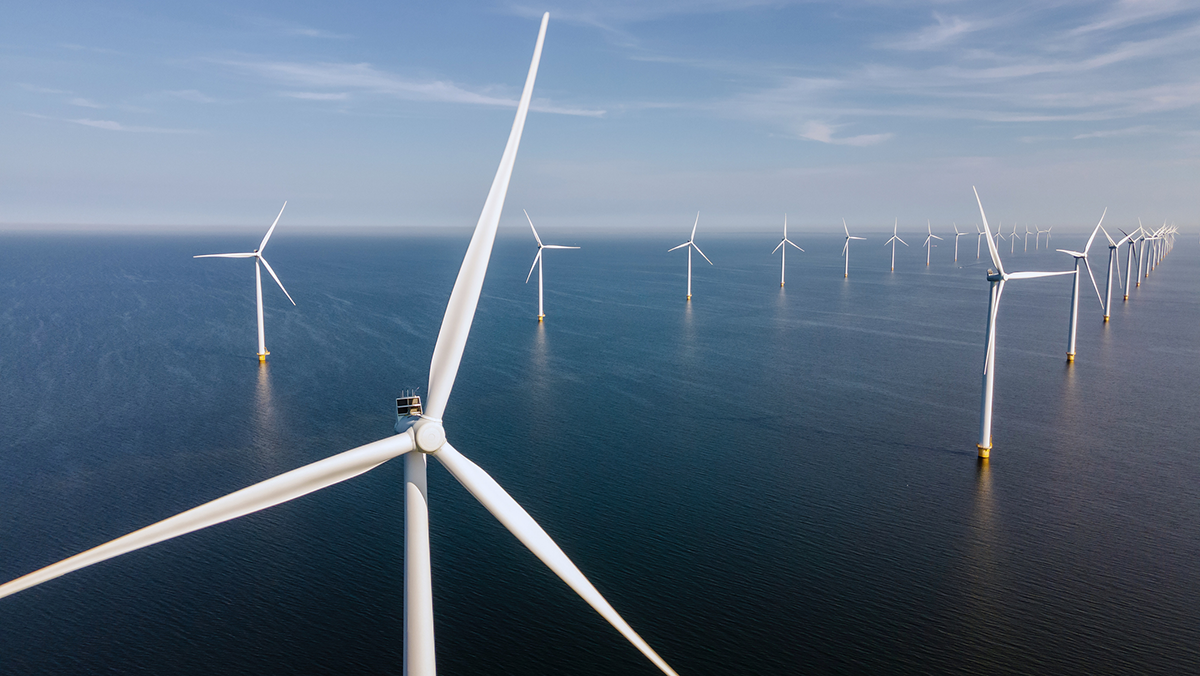 Luchtfoto van een offshore windpark op zee, waar tientallen hoge witte windturbines in lange rijen zijn opgesteld. 
