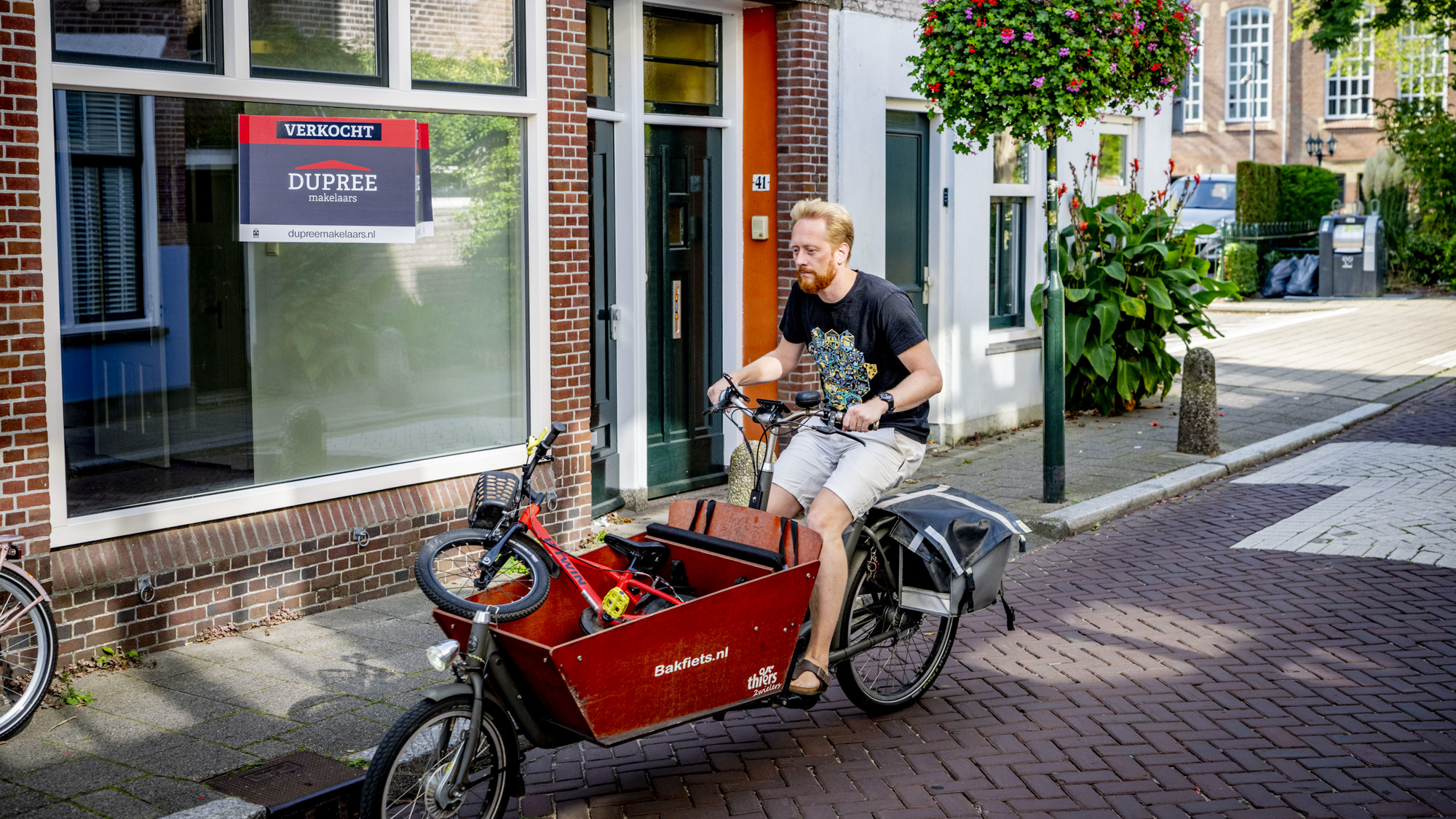 Man fiets met bakfiets langs een huis dat te koop staat.