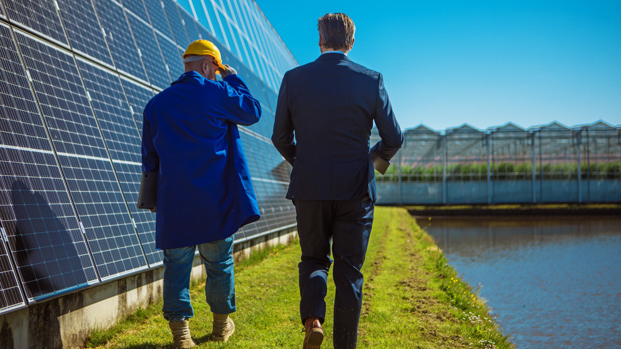 Twee mannelijke ingenieurs lopen langs een rij kassen met zonnepanelen.
