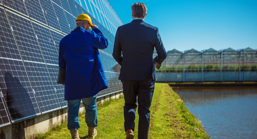 Twee mannelijke ingenieurs lopen langs een rij kassen met zonnepanelen.