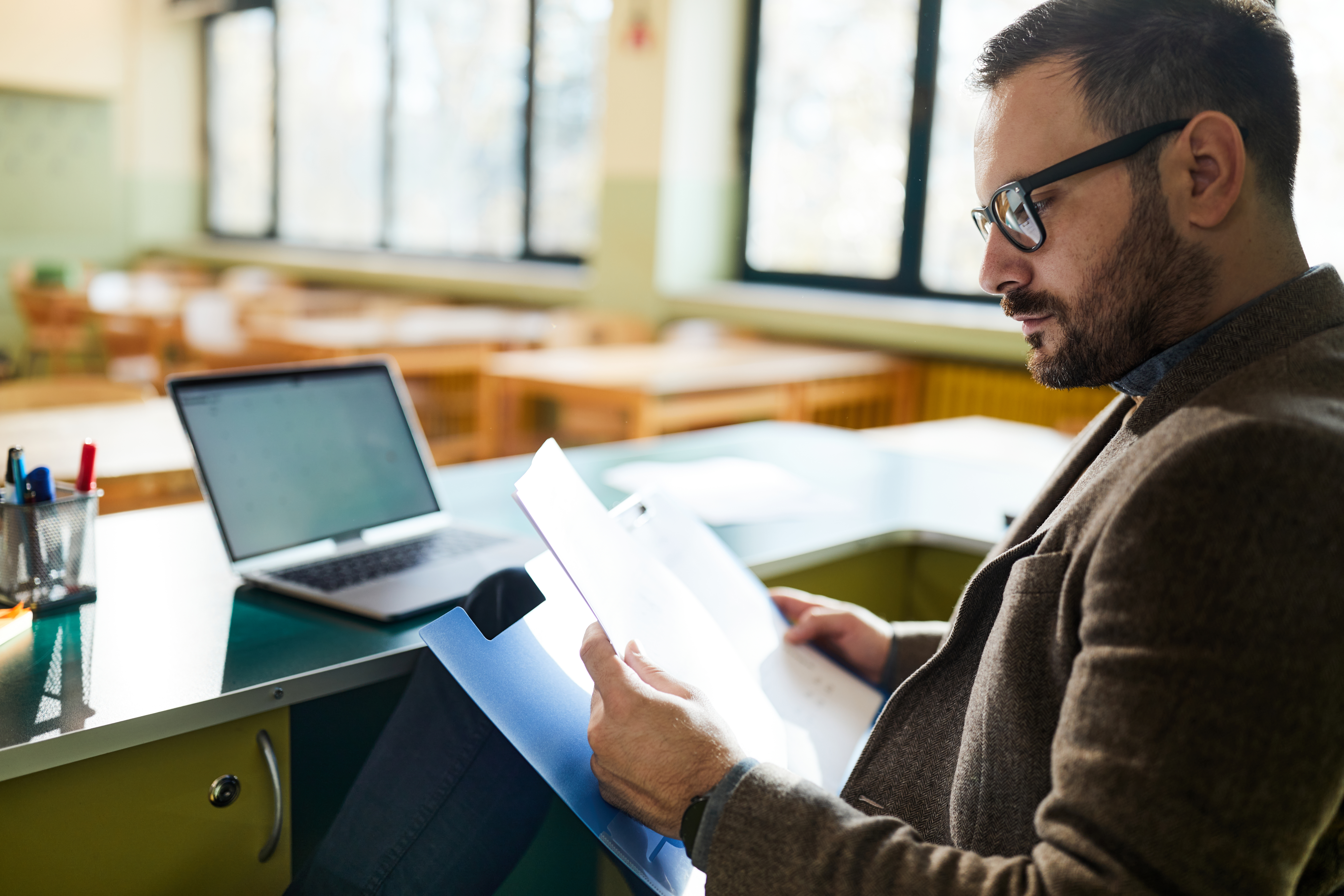 Docent zit aan een bureau en leest documenten, met een open laptop op tafel in een lichte onderwijsomgeving.