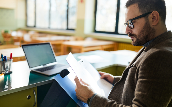 Docent zit aan een bureau en leest documenten, met een open laptop op tafel in een lichte onderwijsomgeving.