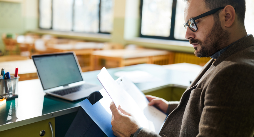 Docent zit aan een bureau en leest documenten, met een open laptop op tafel in een lichte onderwijsomgeving.