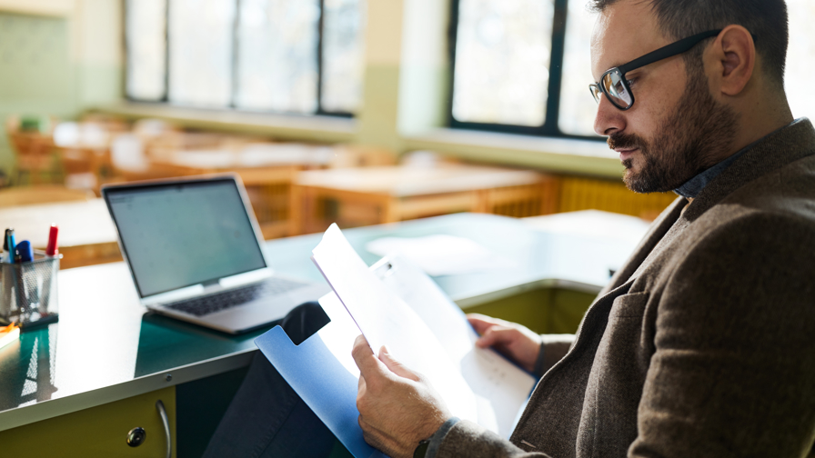 Docent zit aan een bureau en leest documenten, met een open laptop op tafel in een lichte onderwijsomgeving.