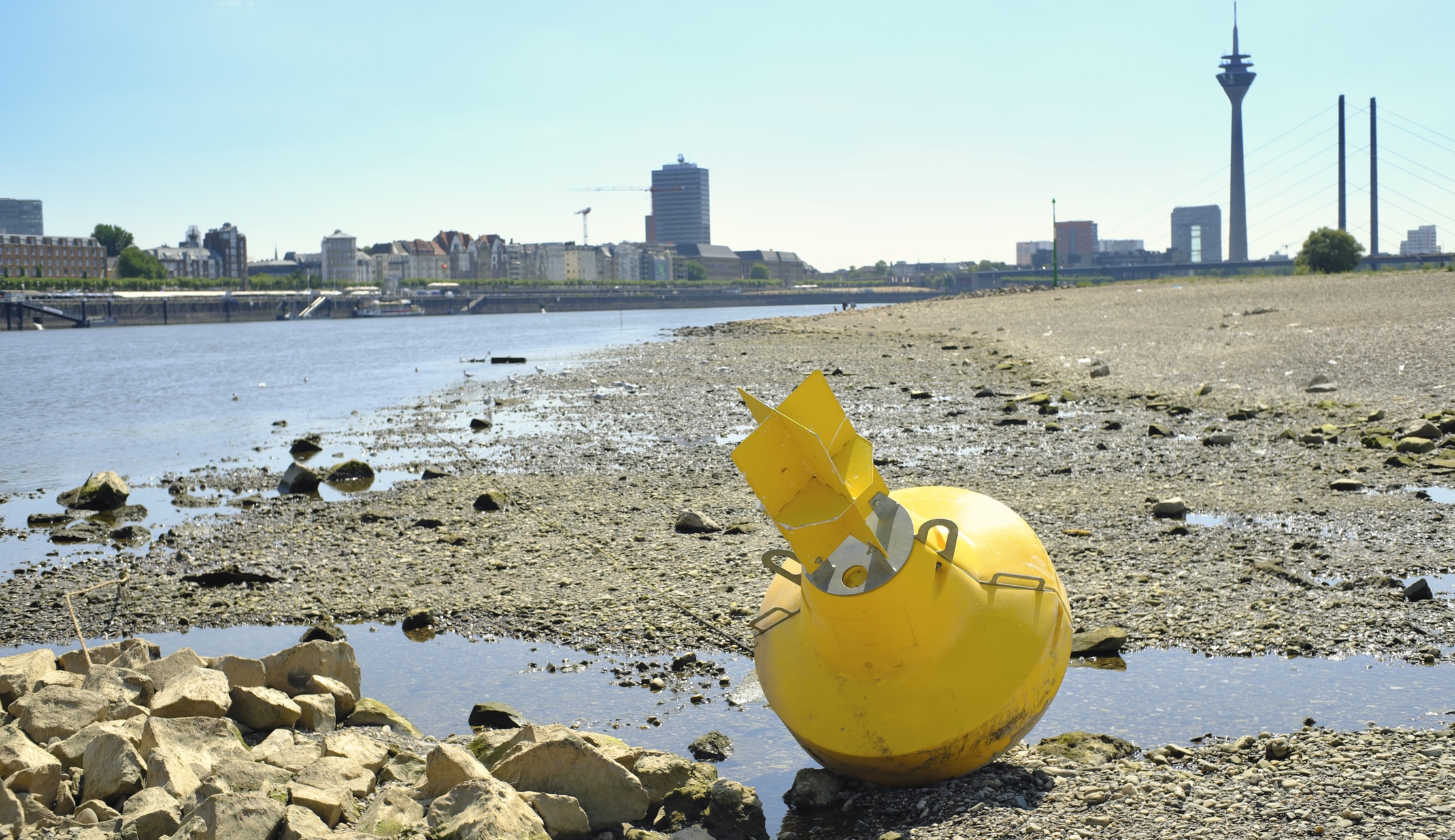 Gele boei op drooggevallen rivieroever langs de Rijn, met de skyline van Düsseldorf op de achtergrond tijdens laagwater.