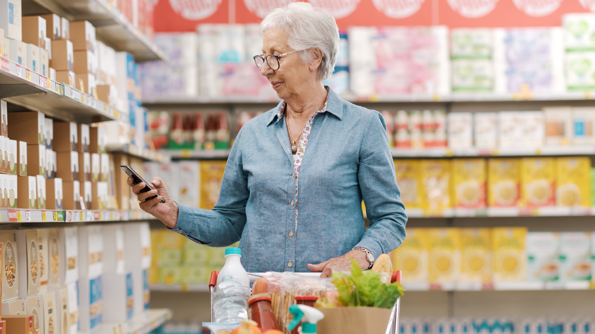 Oudere vrouw checkt prijsstijging door inflatie op haar telefoon tijdens het doen van haar boodschappen in de supermarkt.
