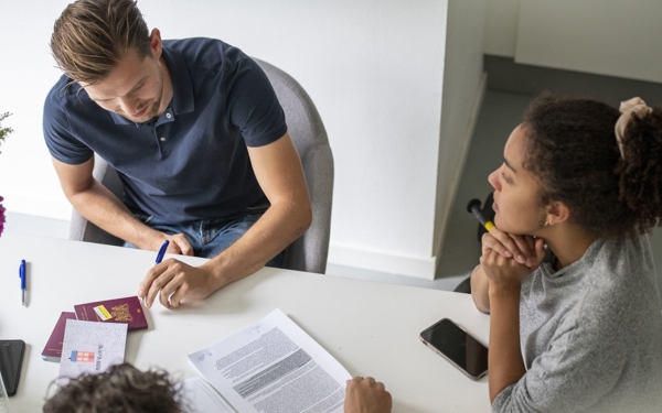 Een stel zit aan een tafel en ondertekent het koopcontract voor hun nieuwe huis. Voor hen liggen verschillende documenten, paspoorten en een identiteitskaart. Aan de andere kant van de tafel zit de notaris, die het papierwerk begeleidt tijdens de officiële overdracht.