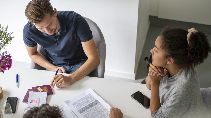 Een stel zit aan een tafel en ondertekent het koopcontract voor hun nieuwe huis. Voor hen liggen verschillende documenten, paspoorten en een identiteitskaart. Aan de andere kant van de tafel zit de notaris, die het papierwerk begeleidt tijdens de officiële overdracht.