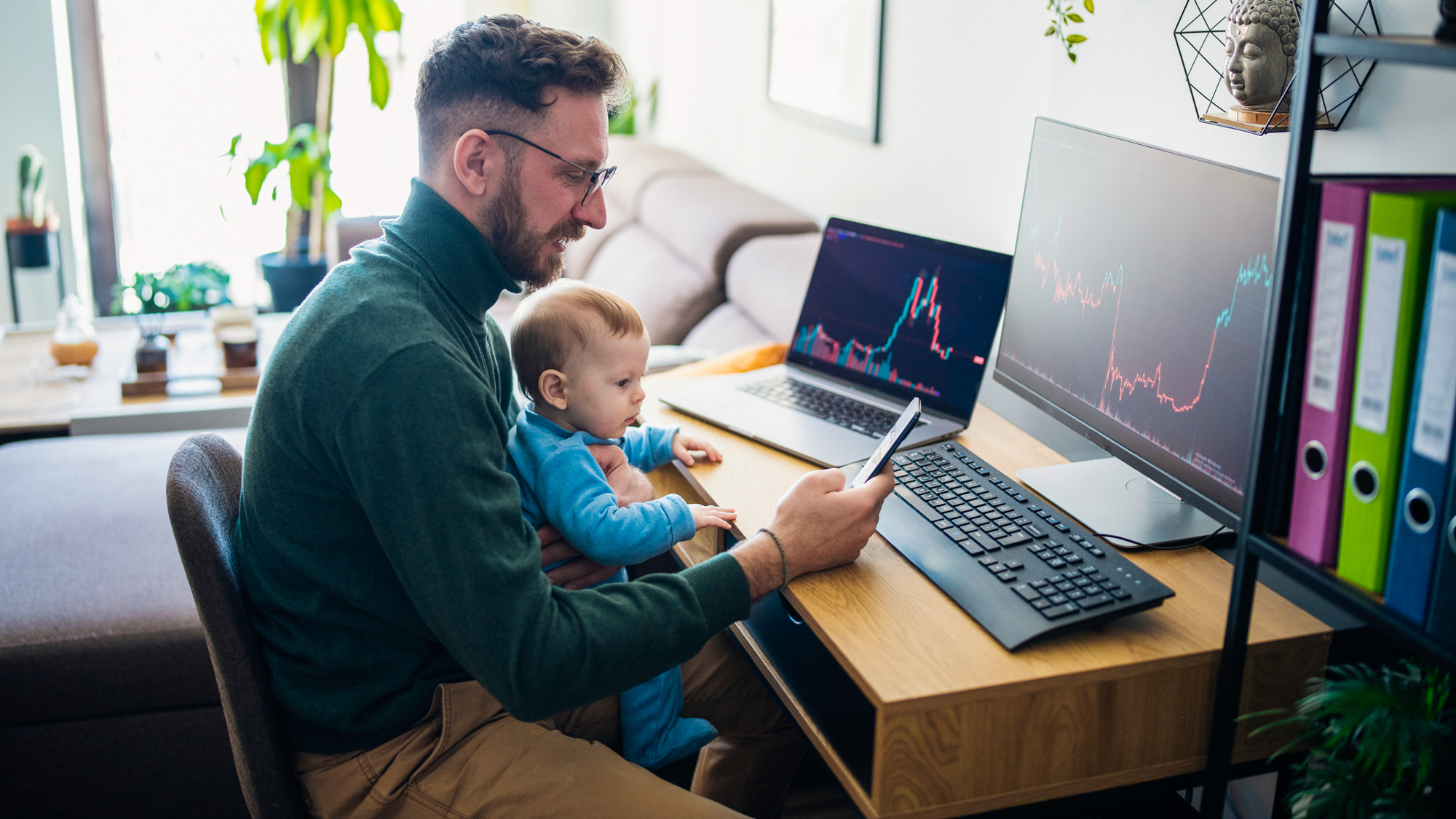 Man met baby op schoot kijkt naar de aandelenmarkt op zijn computer en telefoon.