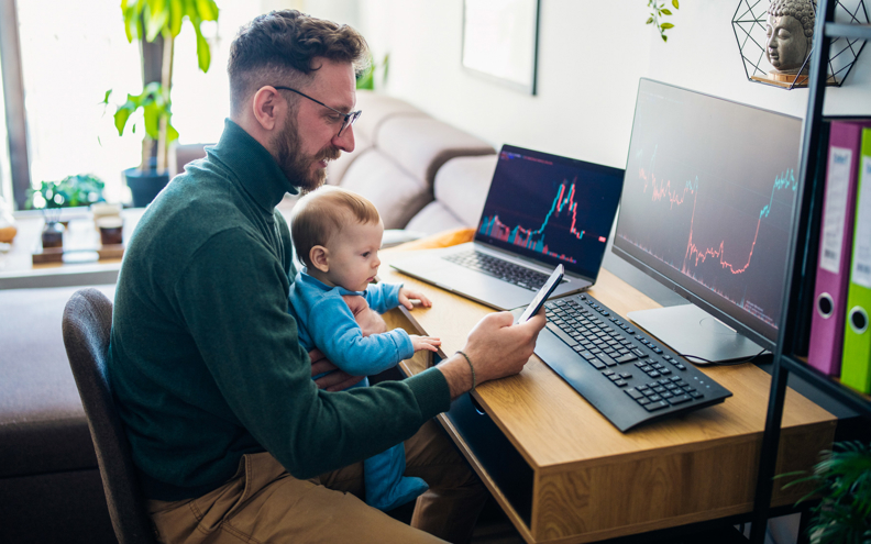Man met baby op schoot kijkt naar de aandelenmarkt op zijn computer en telefoon.