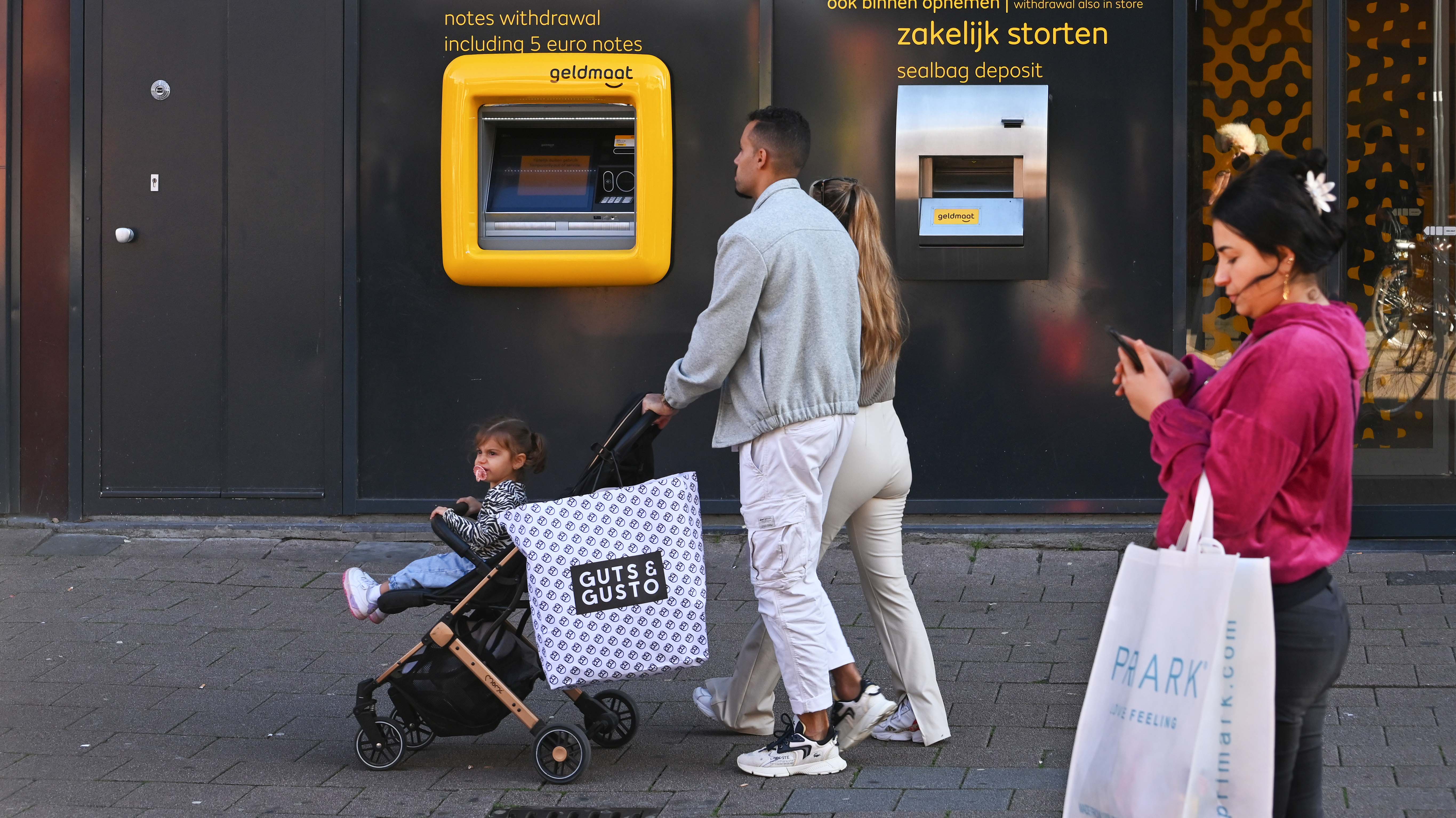 Voor een Geldmaat-geldautomaat lopen winkelende bezoekers over een stoep in een winkelstraat. Op de voorgrond rijdt een kinderwagen met een kind erin, terwijl een grote boodschappentas aan de wagen hangt. Rechts loopt een persoon met een winkeltas en een mobiele telefoon. Op de donkere gevel zijn de gele Geldmaat-automaat en teksten over geld opnemen en zakelijk storten zichtbaar.