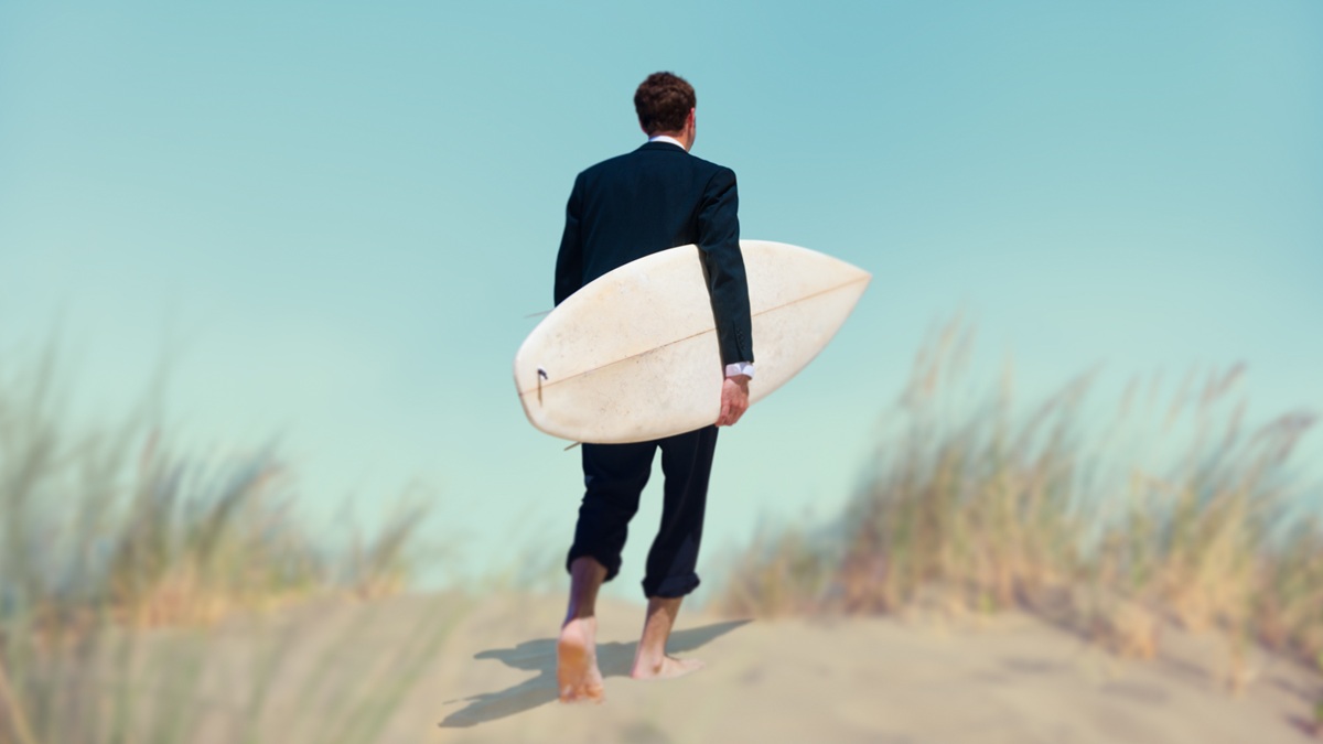 Een man in pak loopt met een surfplank onder zijn armen door een duinlandschap