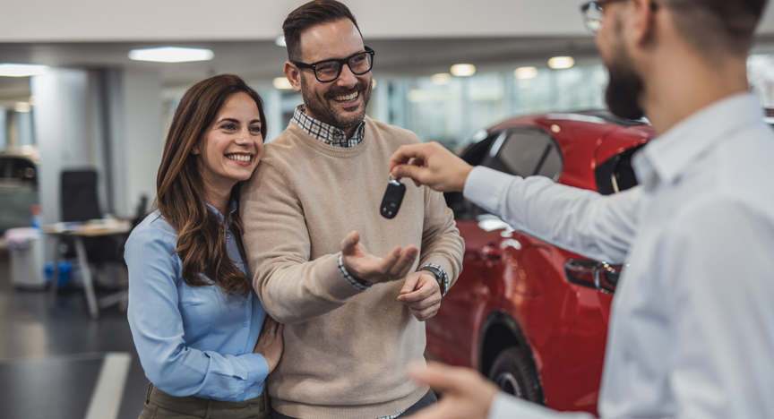 Stel ontvangt de sleutels van hun nieuwe rode auto van een autoverkoper in een showroom.