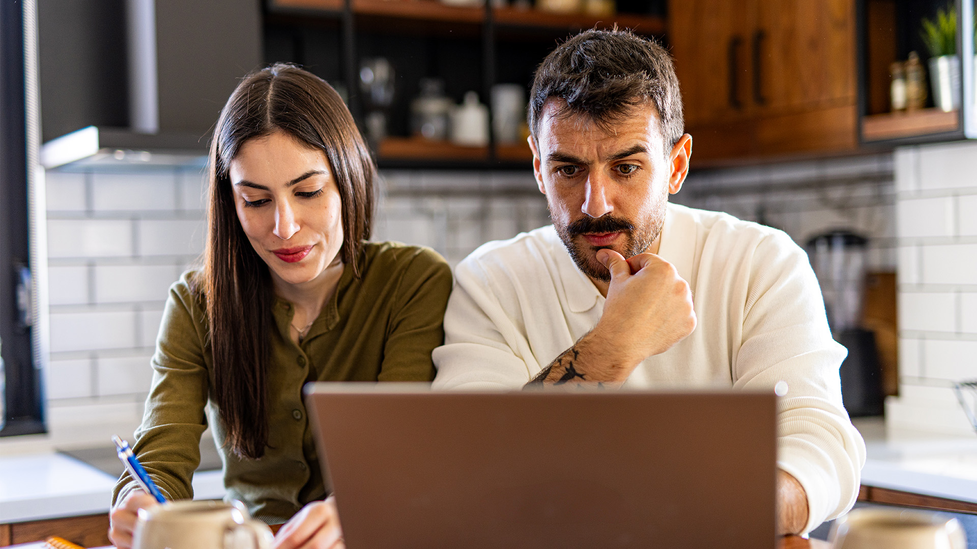 Twee personen zitten samen aan een tafel en kijken naar een laptop in een huiselijke omgeving.